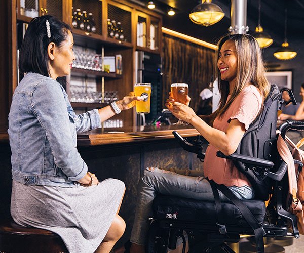 dos mujeres sonriendo y tomando una cerveza en un bar, una de ellas usa silla de ruedas